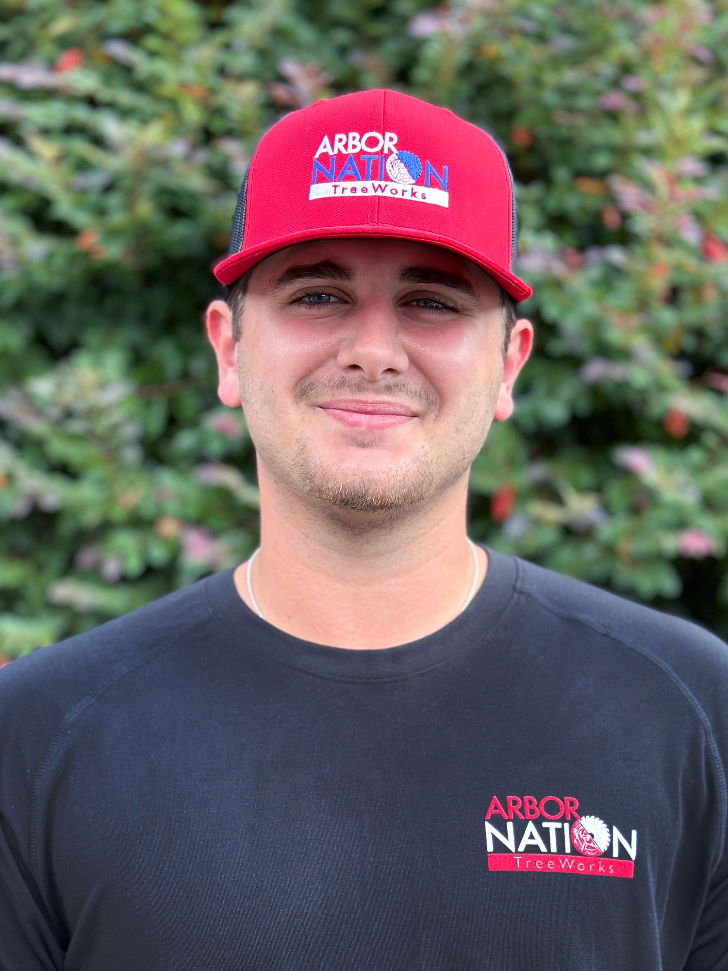 Man wearing a red cap and black t-shirt with Arbor Nation logo against a green bush background