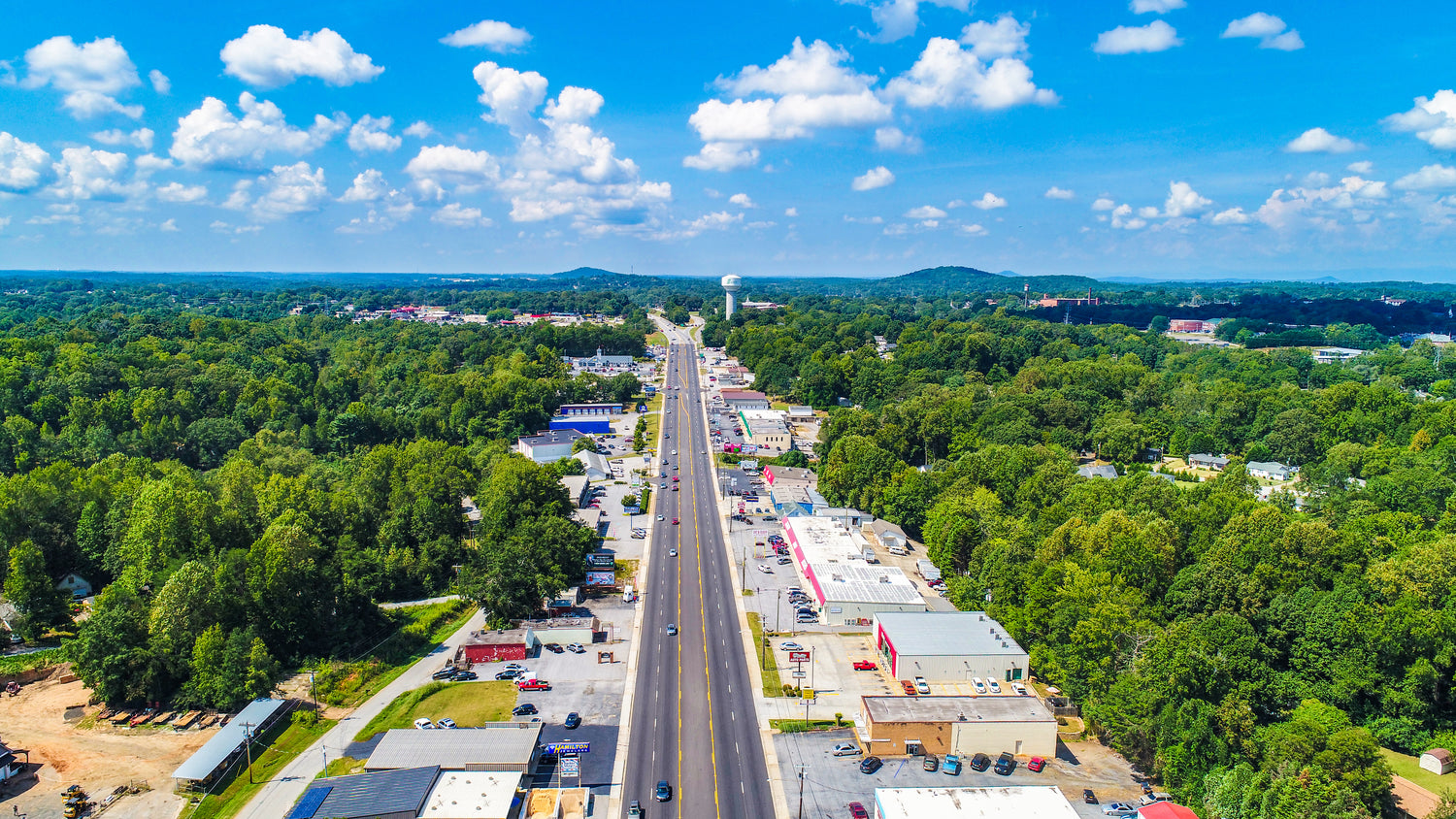 Aerial view of a highway surrounded by green trees under a blue sky with clouds.