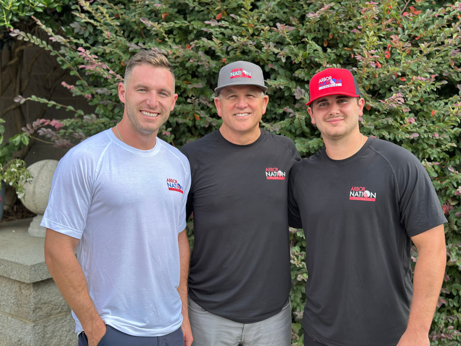 Three men wearing shirts with a logo standing in front of greenery