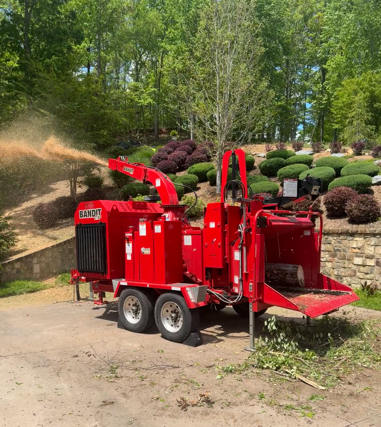 Red wood chipper machine in a garden setting with trees and shrubs in the background.